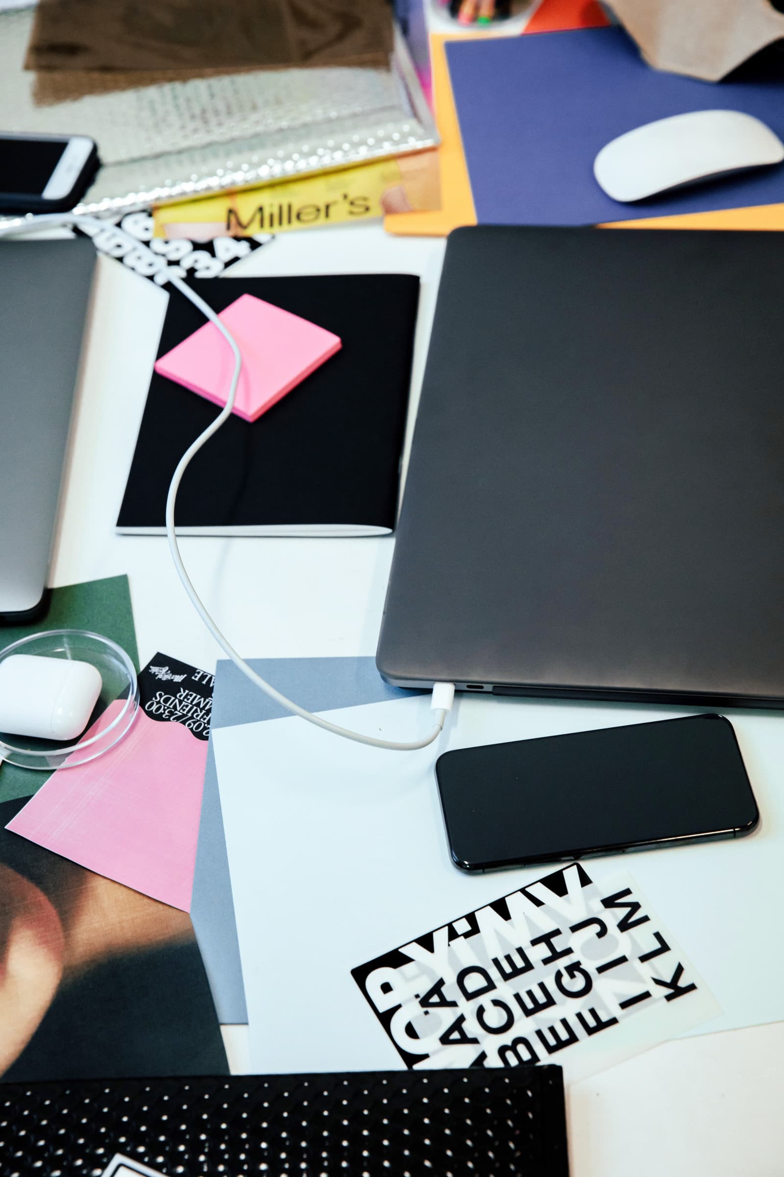 Overhead view of a desk with a closed laptop plugged into a charging cable, a phone, notebook with sticky notes and papers