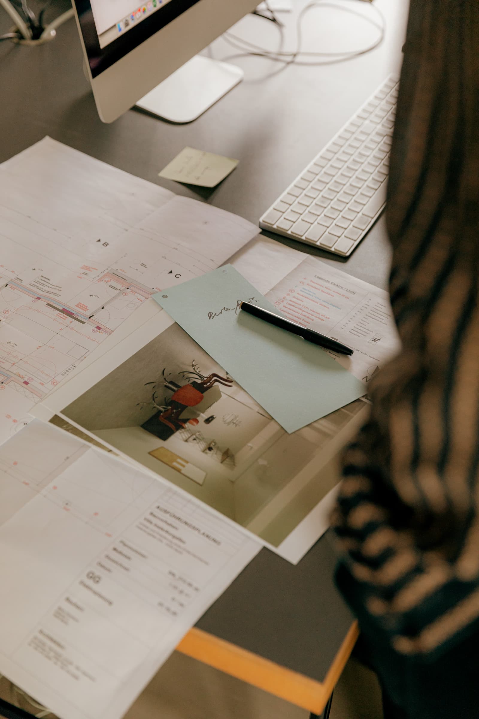 Floor plans, a printed interior photo and a pen spread across a desk beside a keyboard, seen over a person's shoulder
