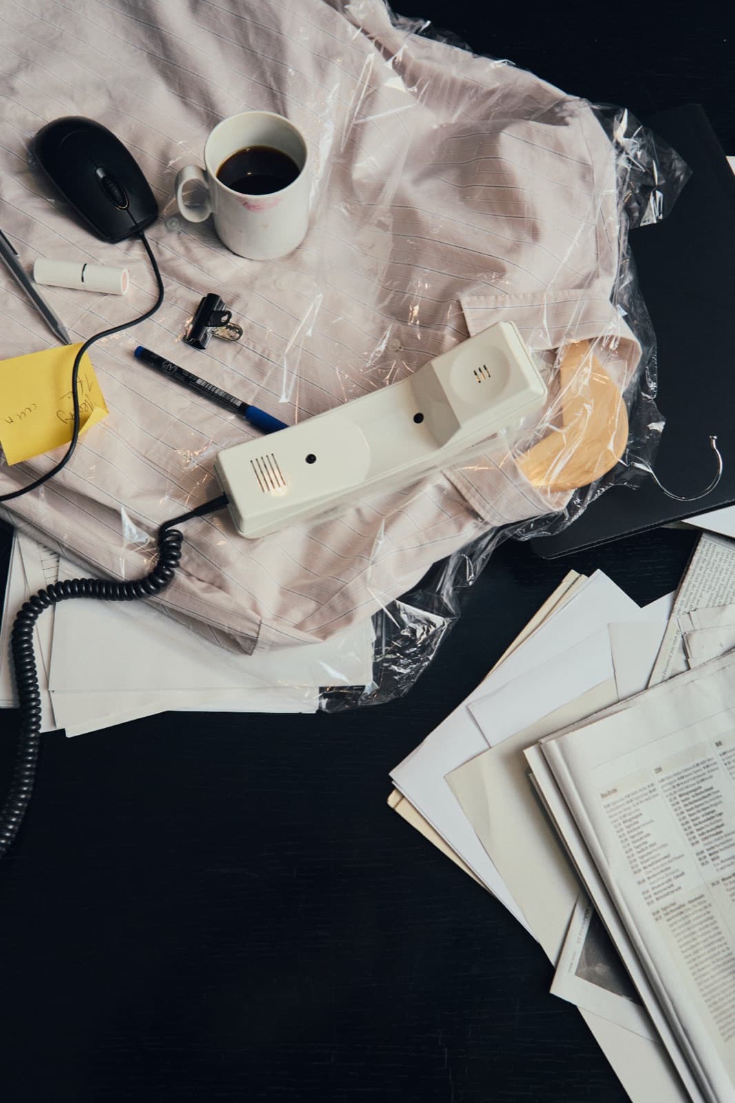 Cluttered desk from above: a corded phone handset, coffee mug and mouse on a plastic-wrapped shirt beside loose papers